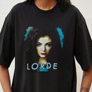 以下が含まれることがあります： 黒いTシャツ。暗い髪と赤い口紅をした女性の肖像画が描かれており、その下に白いブロック体で「LORDE」という名前が書かれています。画像には青色のアクセントがあります。