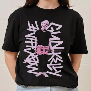 May include: Black t-shirt with a pink graphic of a person holding a pink guitar. The words "MACHINE GUN KELLY" are printed in a graffiti-style font. The person is wearing black clothing and white trainers.