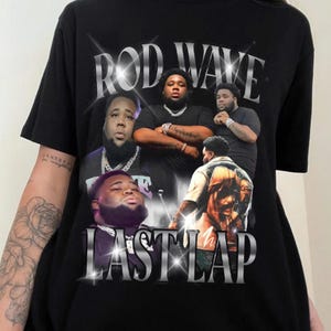 May include: Black t-shirt featuring a graphic design with the text "ROD WAVE" and "LAST LAP" in silver lettering. The design includes multiple images of a man. The shirt is made of a soft material.
