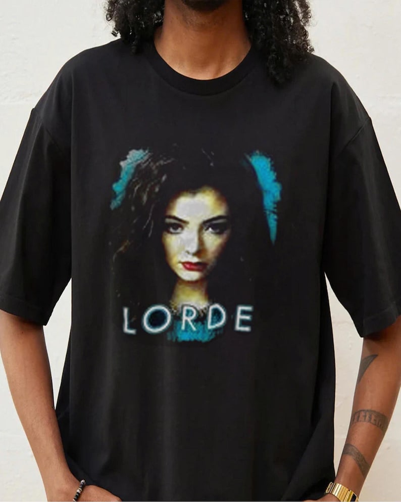 以下が含まれることがあります： 黒いTシャツ。暗い髪と赤い口紅をした女性の肖像画が描かれており、その下に白いブロック体で「LORDE」という名前が書かれています。画像には青色のアクセントがあります。
