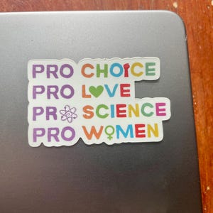 Puede incluir: Una pegatina colorida con las palabras "PRO CHOICE, PRO LOVE, PRO SCIENCE, PRO WOMEN" en varios colores. La pegatina está sobre una superficie gris, con una superficie de madera en el fondo.
