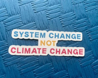 May include: Two white stickers with the text "SYSTEM CHANGE NOT CLIMATE CHANGE" in blue, yellow, and pink lettering. The stickers are on a textured blue surface.