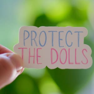 Może przedstawiać: Biała naklejka z napisem "PROTECT THE DOLLS" w niebieskich i różowych literach. Naklejka jest trzymana na tle rozmytego zielonego tła.