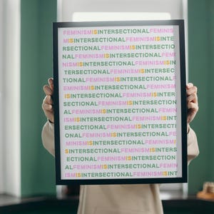 May include: A framed print with the words "FEMINISMISINTERSECTIONALFEMINIS" in a repeating pattern. The text is in various colors, including pink, green, and yellow, against a white background. The frame is black.