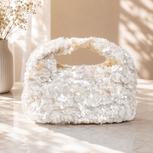 May include: A small, rectangular handbag completely covered in shimmering white sequins. The bag features a structured, integrated top handle and a compact, rounded silhouette, creating a textured, reflective surface that catches the light.