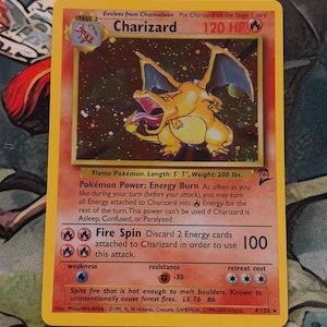 May include: A Charizard Pokémon trading card. The card features a fiery orange Charizard illustration with blue wings, set against a vibrant background. The card text includes the name, HP, and attack details. The card is a Stage 2 evolution.