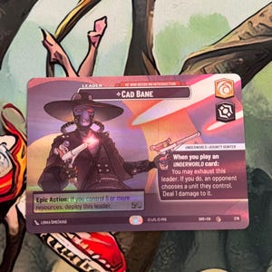 May include: A trading card featuring Cad Bane, a bounty hunter, with a wide-brimmed hat and two blasters. The card has text that reads "Epic Action: If you control 6 or more resources, deploy this leader."