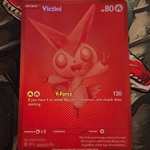 May include: Red Victini Pokémon trading card. The card features a stylized illustration of Victini, with the text "Victini" and "V-Force". The card also includes the HP value, attack power, and other game-related information. The card is from the 2025 Pokémon/Nintendo/Creatures/GAME FREAK collection.