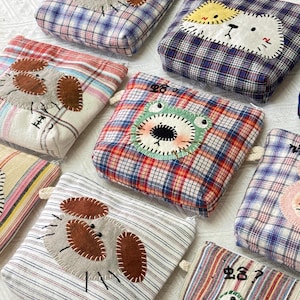 May include: A selection of square fabric pouches, each adorned with an animal face appliqué. The pouches display a variety of plaid and striped patterns in blue, red, and white. Each pouch showcases a different animal design, such as a dog, cat, and frog.