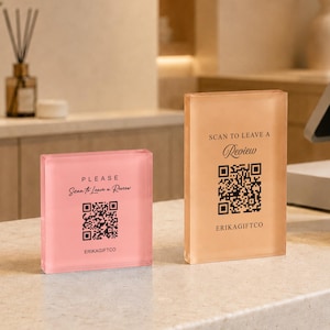 May include: Two rectangular acrylic signs with black text and QR codes. The left sign is pink and reads PLEASE Scan to Leave a Review ERIKAGIFTCO. The right sign is peach-colored and reads SCAN TO LEAVE A Review ERIKAGIFTCO. Both are upright.