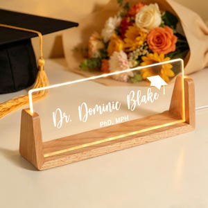 May include: A wooden desk nameplate with the text "Dr. Dominic Blake, PhD, MPH" illuminated by a warm light. A graduation cap with a gold tassel is in the background, along with a bouquet of flowers. The nameplate is a graduation gift.
