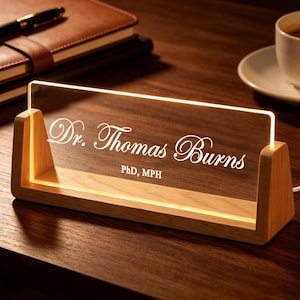 May include: A lighted desk nameplate with the text "Dr. Thomas Burns, PhD, MPH" in white script. The nameplate is made of clear acrylic and sits in a wooden base. A pen, notebook, and coffee cup are in the background.