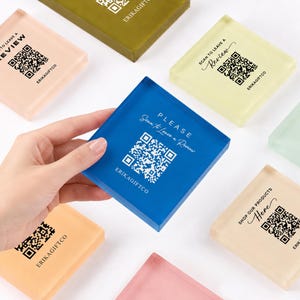 Può includere: Un cartello quadrato in acrilico blu con un codice QR bianco, il testo "PLEASE Scan to Leave a Review" e "ERIKAGIFTCO" stampati sulla superficie. Il cartello è tenuto davanti ad altri cartelli quadrati in acrilico di vari colori pastello.