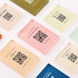 Può includere: Una collezione di blocchi rettangolari in acrilico in vari colori pastello, ciascuno con un codice QR stampato e del testo. Il testo include frasi come "PLEASE Scan to Leave a Review", "SCAN TO LEAVE A Review" e "SHOP OUR PRODUCTS Here", con "ERIKAGIFTCO" stampato sotto ogni codice QR.