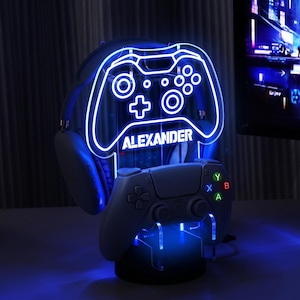 Può includere: Un supporto da gaming in acrilico trasparente con un contorno LED blu luminoso a forma di controller di gioco, con il nome ALEXANDER stampato al centro. Il supporto sostiene un paio di cuffie scure e un controller wireless con tasti colorati.