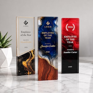 Puede incluir: Tres premios rectangulares de acrílico con el texto Employee of the Year. El primero es blanco con patrones de mármol negro y dorado. El segundo es de mármol azul y blanco con base de madera. El tercero es rojo y negro con una corona de laurel 2026.