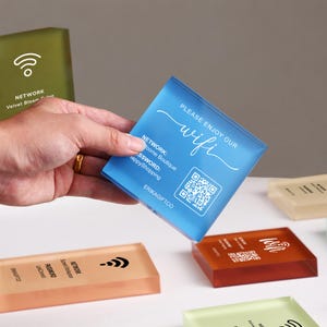 May include: A square blue acrylic sign featuring the white text PLEASE ENJOY OUR wifi, NETWORK: Welcome Boutique, PASSWORD: HappyShopping, a QR code, and the brand name ERIKAGIFTCO. Other translucent colored acrylic signs are visible nearby.