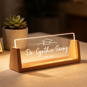 Könnte beinhalten: Ein beleuchtetes Namensschild für den Schreibtisch mit Holzsockel und klarer Acrylplatte. Auf der Platte steht "Dr. Cynthia Saenz, PHD, MPH" und eine Abschlusskappe. Das Schild leuchtet warm.