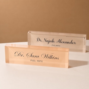 May include: Two rectangular clear acrylic desk nameplates. The front plate is peach-tinted with black text reading Dr. Sara Wilkins PhD, MPH. The back plate is clear with black text reading Dr. Najah Alexander PhD, MPH in a script font.