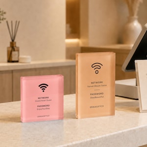 May include: Two rectangular acrylic signs with black text and Wi-Fi icons. The smaller pink sign displays NETWORK Grand Hotel Guest, PASSWORD EnjoyYourStay, and ERIKAGIFTCO. The taller peach sign displays NETWORK Velvet Bloom Salon, PASSWORD StayBeautiful, and ERIKAGIFTCO.