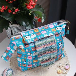 May include: A turquoise fabric project bag with a zipper closure, adorned with a repeating pattern of cartoon animals. A partially knitted sock rests on the bag. A ball of yarn and a small doily are also visible.