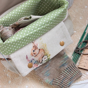 Può includere: Un cestino foderato in tessuto a pois verdi e bianchi contiene filato e una piccola pochette con un'illustrazione di un coniglio. Sono visibili anche un calzino lavorato a maglia e ferri da maglia. Il cestino è su una superficie bianca.