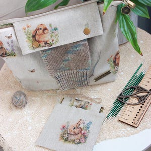 Può includere: Una borsa tote e una piccola pochette in lino beige presentano un'illustrazione ad acquerello di un coniglietto in un giardino fiorito. Sono presenti anche un calzino lavorato a maglia, un gomitolo di lana, ferri da maglia, forbici e un righello. La borsa ha una cerniera e una patta con un bottone.