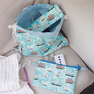 Può includere: Un sacco da progetto azzurro con coulisse e una pochette con cerniera abbinata, entrambi decorati con un motivo di barche a vela nautiche. Sono visibili anche un pezzo all'uncinetto bianco, un uncinetto viola e delle forbici.