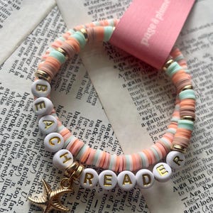 May include: A double-strand bracelet made of small, flat, circular clay beads in shades of peach, mint green, and tan, accented with gold-tone spacers. White round beads spell out BEACH READER in gold letters, and a gold-tone starfish charm hangs from the side.