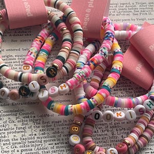 May include: A collection of stretch bracelets made from colorful polymer clay heishi beads. The bracelets feature gold-toned accents and white letter beads that spell out the word BOOKISH. Each bracelet is attached to a pink paper tag labeled page & plume.