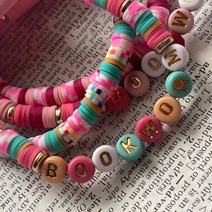 May include: Stack of colorful beaded bracelets with the word "BOOKWORM" spelled out in gold letters. The bracelets feature pink, red, teal, and white disc beads. The background is a page from a dictionary.