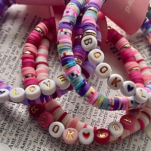 May include: A collection of stacked bracelets made from colorful polymer clay heishi beads in shades of pink, purple, and blue, accented with gold-tone spacers and white round beads featuring gold letters that spell out the words BOOK LOVER.