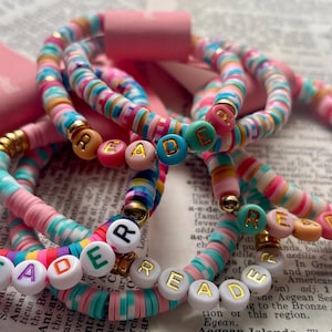 May include: Colorful beaded bracelets with the word "READ" spelled out in letter beads. The bracelets feature a mix of pink, blue, and gold accents. The bracelets are stacked together, creating a vibrant and eye-catching accessory.