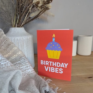 Op de afbeelding: Een verjaardagskaart met een rode achtergrond, met een cupcake-illustratie met een blauwe bovenkant, gele basis en een brandende kaars. De woorden "BIRTHDAY VIBES" staan in witte tekst. De kaart ligt op een houten oppervlak.