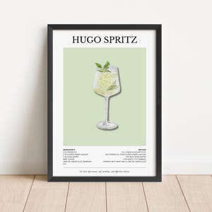 Op de afbeelding: Een ingelijste print met een illustratie van de Hugo Spritz cocktail. Het kunstwerk toont een wijnglas gevuld met de drank, gegarneerd met munt en limoen. De titel "Hugo Spritz" staat bovenaan, met ingrediënten en methode eronder.