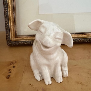 Könnte beinhalten: Eine weiße Keramikfigur einer lächelnden Kreatur mit großen Ohren und geschlossenen Augen. Die Figur sitzt auf einer Holzoberfläche, mit einem gerahmten Kunstwerk im Hintergrund.