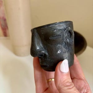 Op de afbeelding: Kleine, donkergrijze keramische sculptuur van een menselijk gezicht. Het gezicht heeft gesloten ogen, een prominente neus en een licht geopende mond. De sculptuur wordt in een hand gehouden, met een witte nagel zichtbaar. Een uniek kunstwerk.