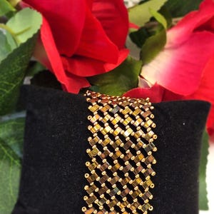 Könnte beinhalten: Ein breites Armband aus einem gewebten Gitter aus metallisch goldenen und schillernd bronzefarbenen rechteckigen Perlen, akzentuiert durch kleine runde goldene Perlen an jeder Kreuzung, was ein strukturiertes, sich wiederholendes geometrisches Muster erzeugt.