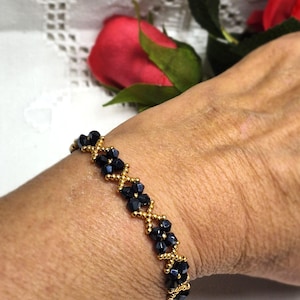 May include: A gold-toned bracelet with a repeating pattern of dark blue beads. The beads are faceted and arranged in a diamond-like design, connected by small gold-colored beads. The bracelet is on a wrist.