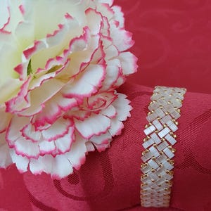 Puede incluir: Una pulsera de cuentas con un diseño tejido, con cuentas rectangulares transparentes y detalles dorados. La pulsera se muestra sobre una superficie roja junto a una flor blanca y rosa. La pulsera mide aproximadamente 2 cm de ancho.