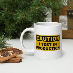 Puede incluir: Una taza de cerámica blanca con un gráfico de señal de advertencia rectangular amarilla con borde negro. El texto en la señal dice CAUTION I TEST IN PRODUCTION en letras mayúsculas negras en negrita. La taza tiene un asa curva.