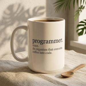 May include: A white ceramic mug with a black printed definition that reads: programmer. noun. An organism that converts coffee into code. The mug features a large handle and is filled with dark coffee, accompanied by a small wooden spoon.