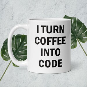 Puede incluir: Taza de café de cerámica blanca con el texto "I TURN COFFEE INTO CODE" en letras negras en negrita. La taza tiene un asa curva y está colocada sobre un fondo gris claro con hojas verdes.
