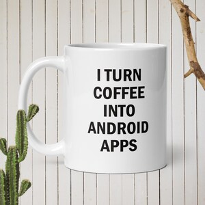 Peut inclure: Mug en céramique blanche avec l'inscription noire "I TURN COFFEE INTO ANDROID APPS". La tasse a une anse incurvée et est posée sur un fond en bois blanc. Un cactus et une branche sont visibles.