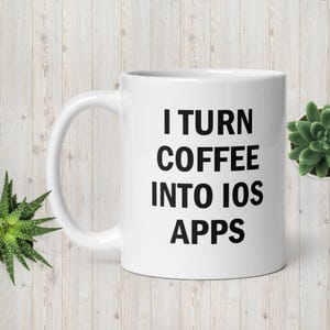 May include: White ceramic mug with the text "I TURN COFFEE INTO IOS APPS" in black. The mug is on a light wood-grain surface, with a small succulent plant in the background. The mug has a curved handle.