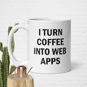 May include: White ceramic mug with the text "I TURN COFFEE INTO WEB APPS" in black. The mug is on a wooden surface with a small copper watering can and a cactus in the background.