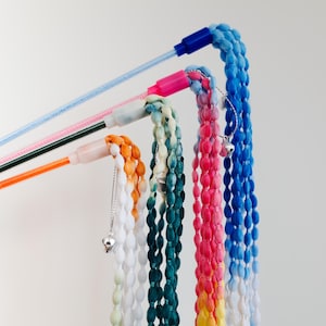 Puede incluir: Una colección de coloridos juguetes para gatos con cuentas. Cada juguete presenta una combinación de colores diferente, incluyendo naranja, blanco, verde azulado, rosa, amarillo y azul. Los juguetes están unidos a varitas finas y coloridas, y algunos tienen pequeños dijes de corazón plateado.