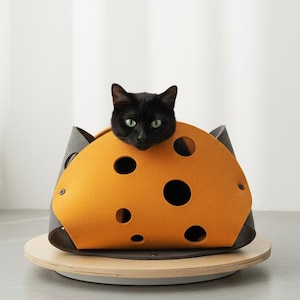 Puede incluir: Un gato negro descansando dentro de una cama para mascotas de fieltro semicircular. La cama presenta una capa exterior de color naranja brillante con recortes circulares que recuerdan al queso suizo, fijada con remaches a un forro gris oscuro.