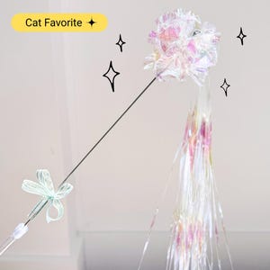 May include: A cat toy with a shimmering, iridescent flower-shaped head and long, flowing streamers. The toy has a thin, black handle and a small bow near the base. The text "Cat Favorite" is in a yellow box.