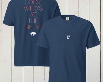 May include: Navy blue t-shirt with the number 17 in white on the front. The back of the shirt features the text "LOOK WHO'S AT THE HELM" in pink, with a white bison graphic below. The shirt is made of a soft material.